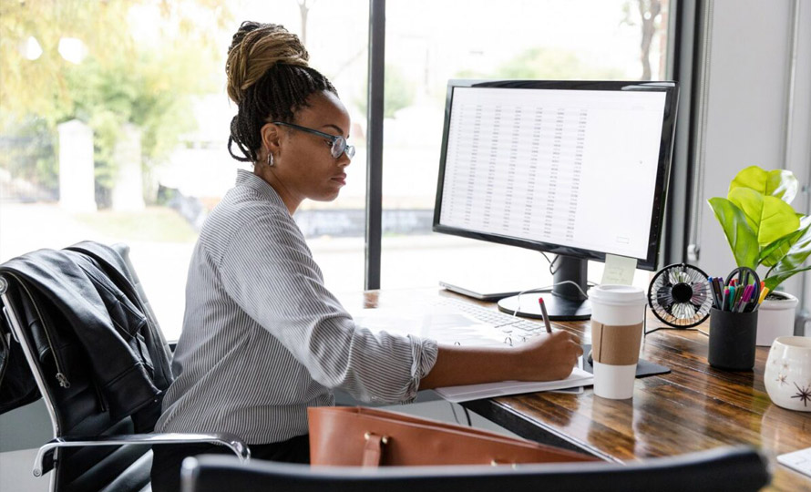 Professional writing at a desk with a computer monitor in a bright office environment.