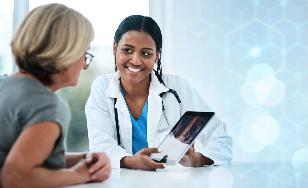 Smiling female doctor in white coat and blue scrubs consulting with a blonde female patient, holding a tablet in a bright medical office