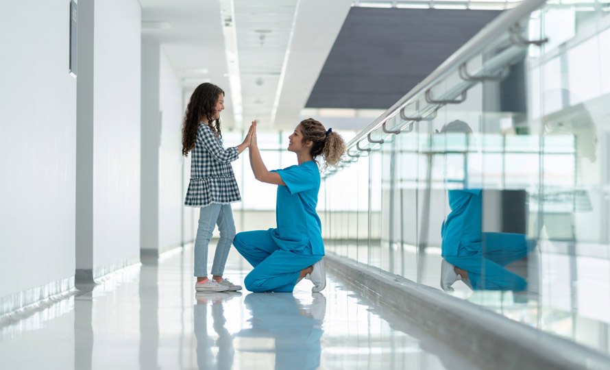 Nurse kneeling to give a high-five to a young girl in a hospital hallway.