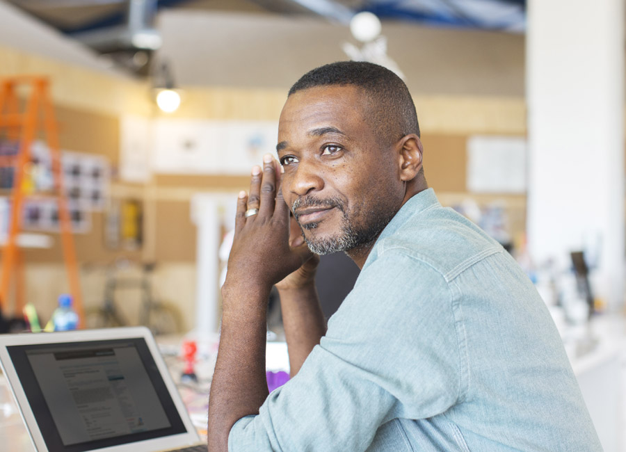 Man in a light denim shirt looking thoughtful while seated at a desk.
