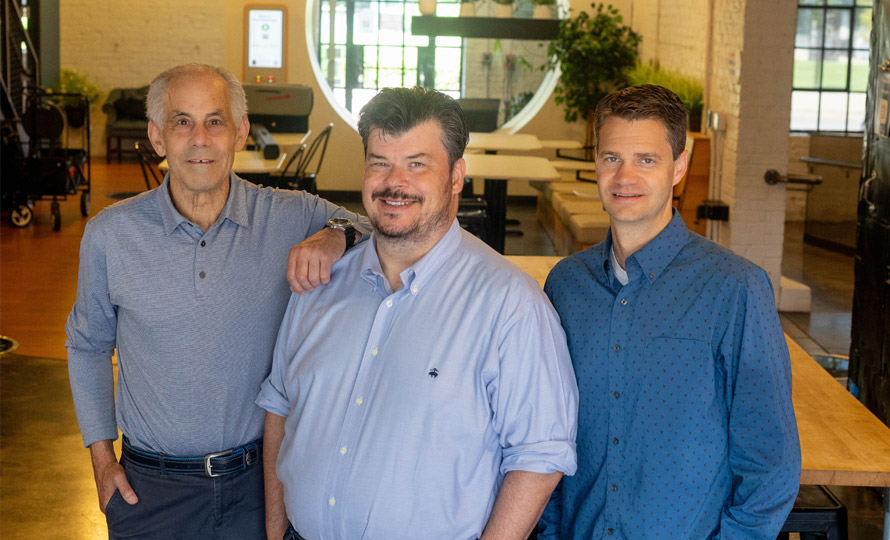 From left to right: Gary Wald (COO), Ben Newton (CEO), and John Clark (CTO) standing together and smiling in an office setting.