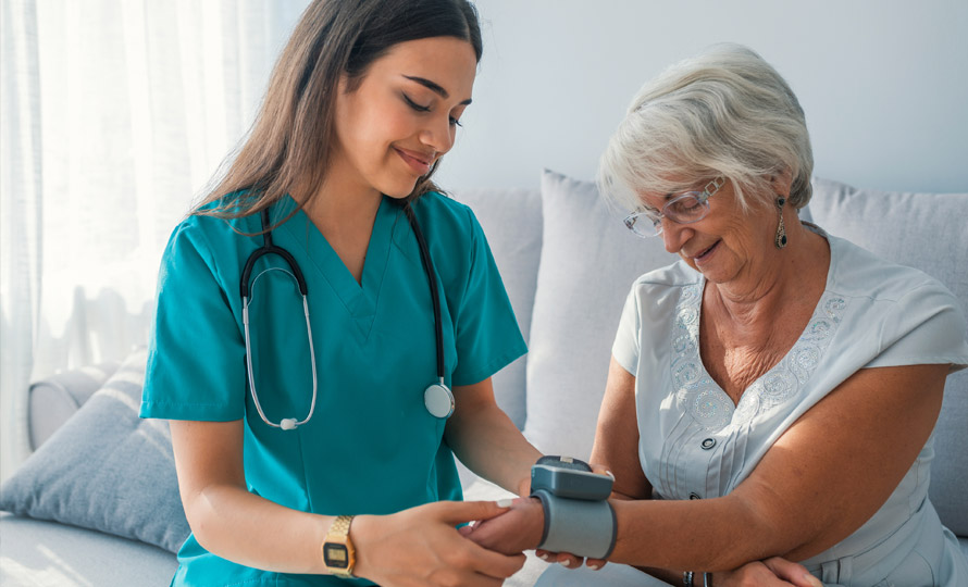Nurse in teal scrubs taking a blood pressure reading for an older woman at home.