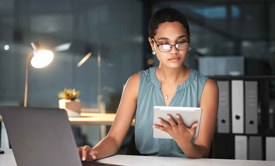 Professional woman working late at a desk, reviewing information on a tablet beside her laptop.