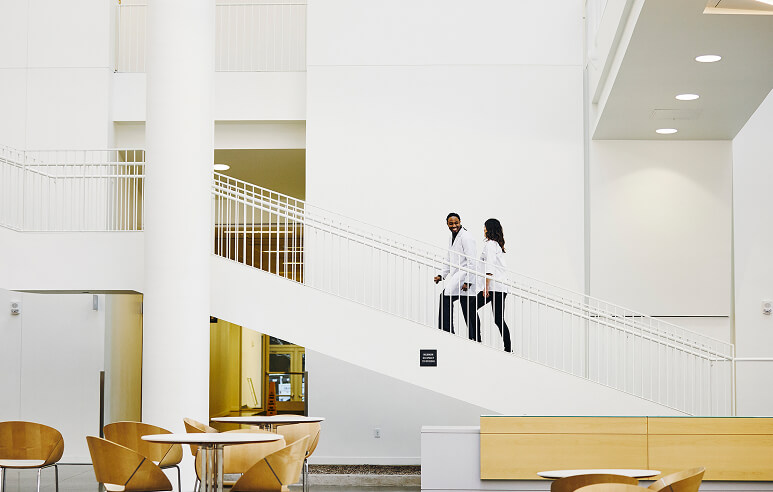 Two medical professionals in white coats walk up a flight of stairs in an office lobby.